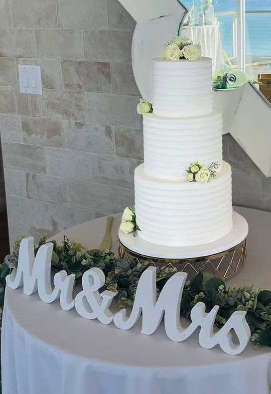 Three-tier white wedding cake with delicate green floral accents and rustic wedding display by Sweet Cakes by Kristen